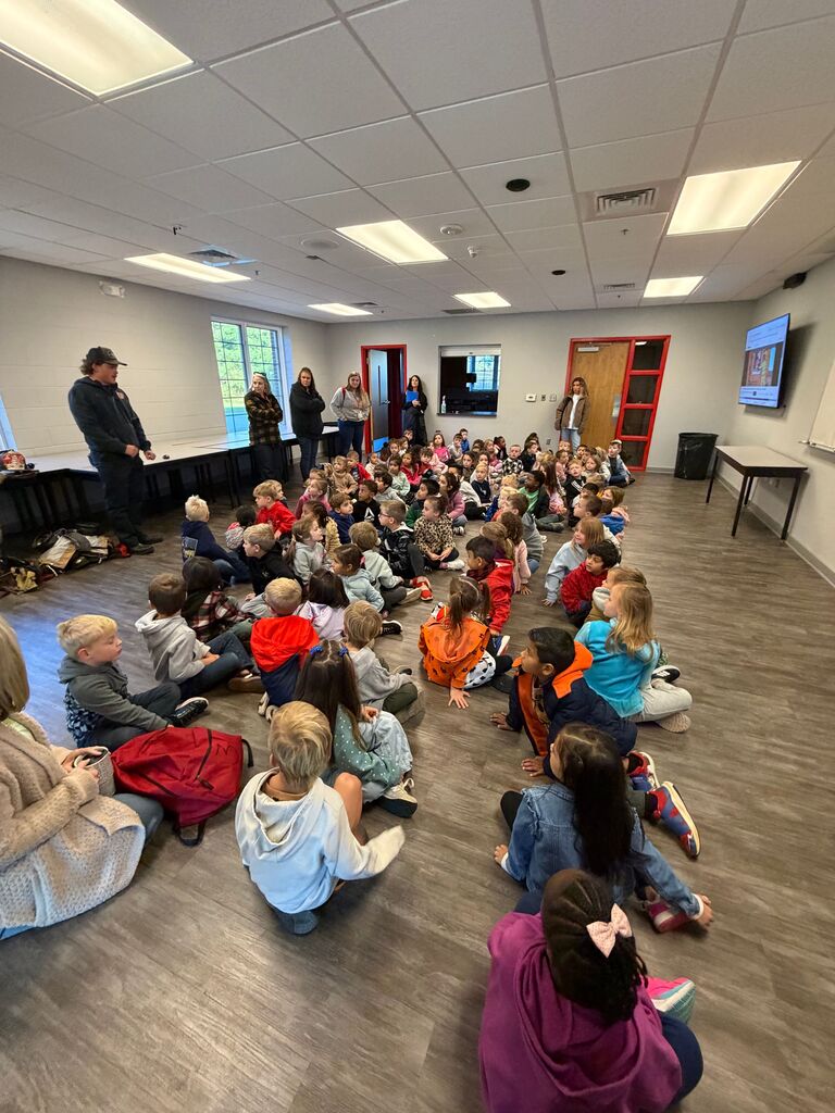 Many children sit on floor, facing man at front of room. Man stands near desk. Room has large windows, wooden floor, and white walls.