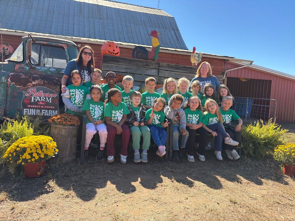 Group of children in green shirts posing in front of building with pumpkins, farm truck, and plants.