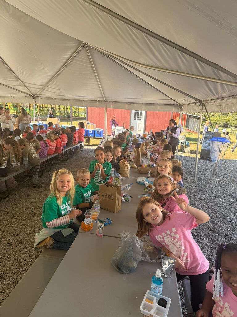 Children sit under a tent at a long table, eating food from paper bags. Some wear green shirts.