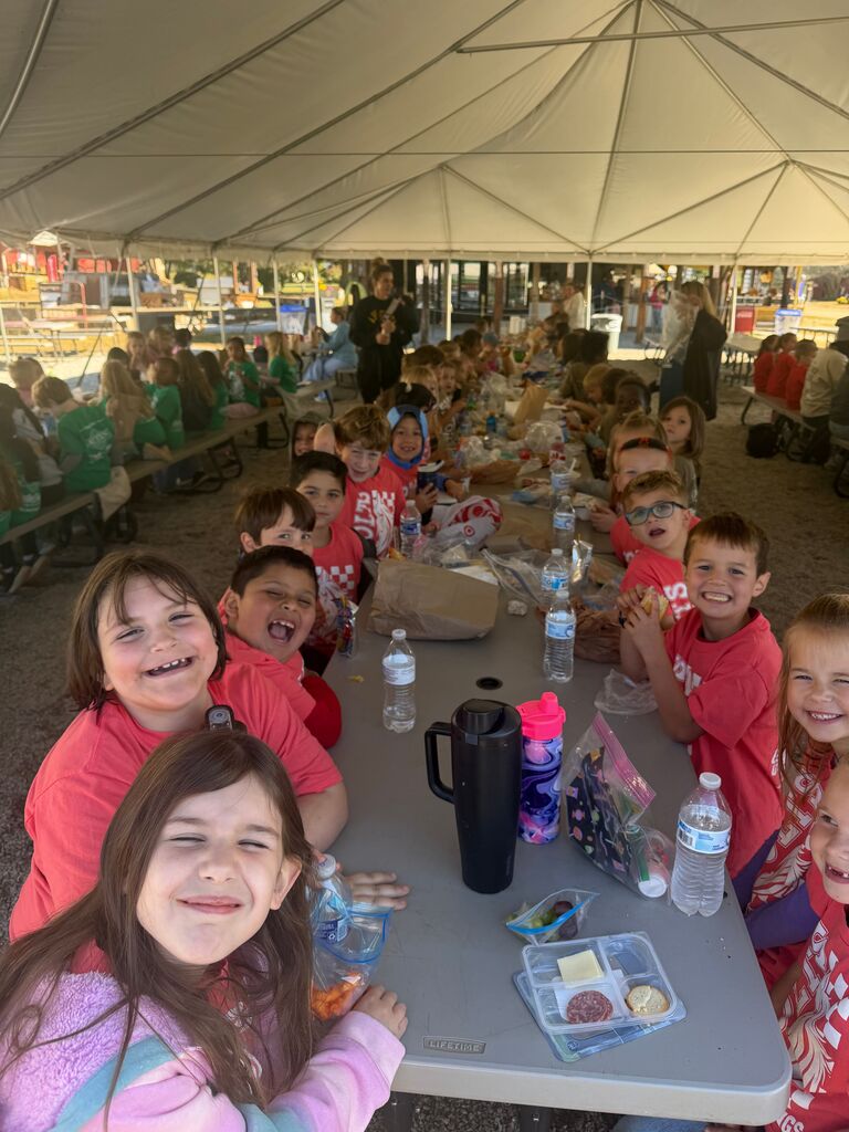Group of children in matching red shirts seated at a picnic table with water bottles and food, under a tent.