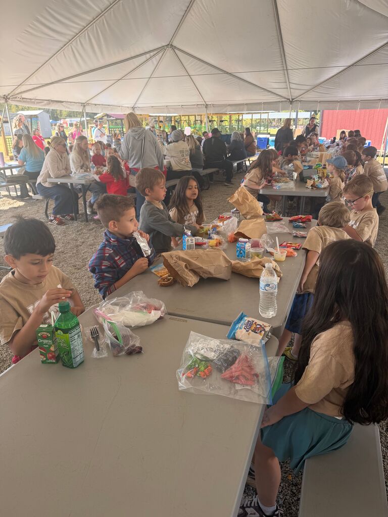 Children eat at tables under a white tent; some have lunches, drinks, and backpacks nearby.