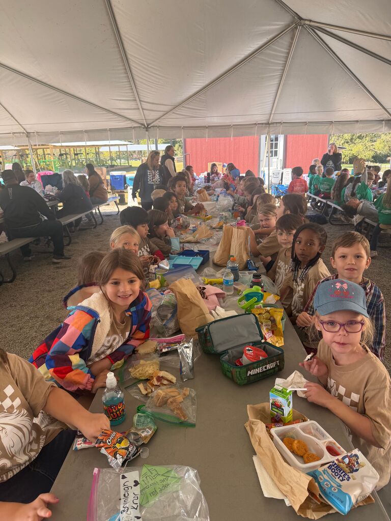 Children sit at a long table under a tent, with trays of food, cups, and bags.