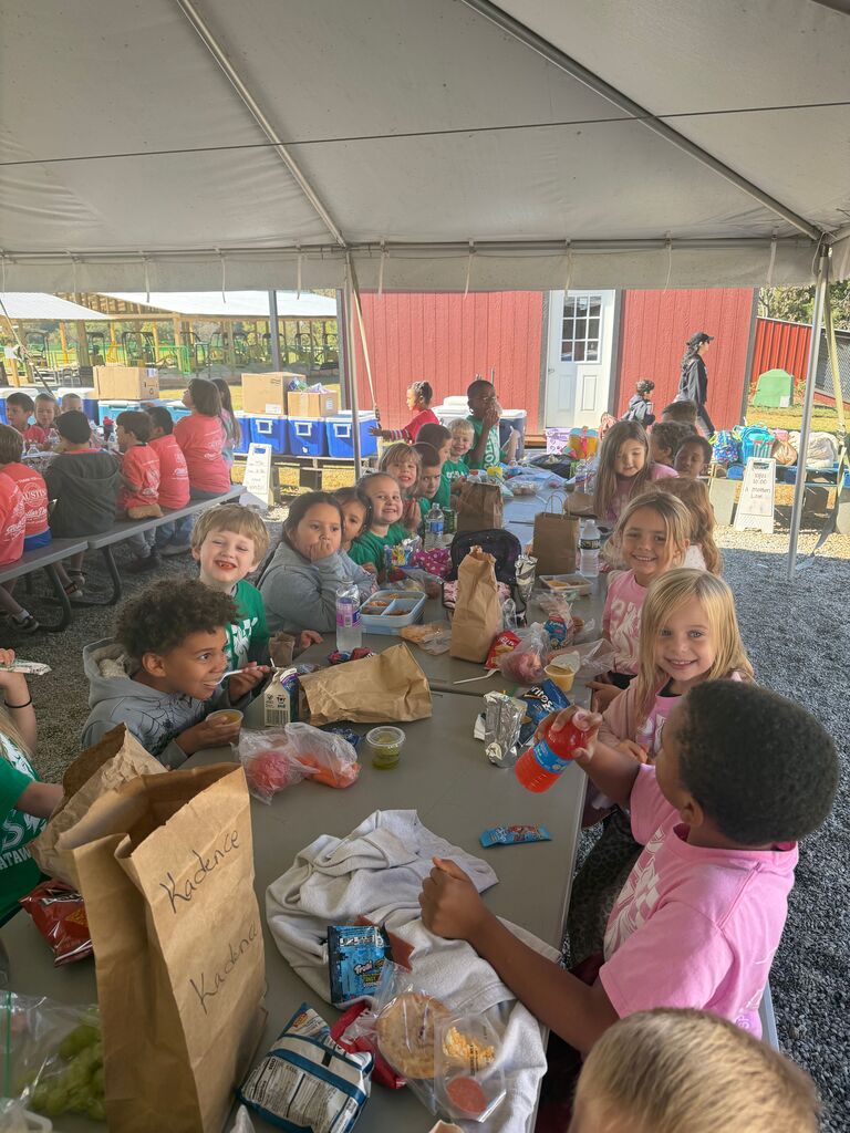 Children seated at a long table under a tent, eating. Brown paper bags, plastic water bottles, and other food items on the table.