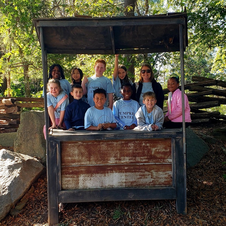 A group of children and adults stand behind a wooden structure in a forest. Some are smiling.