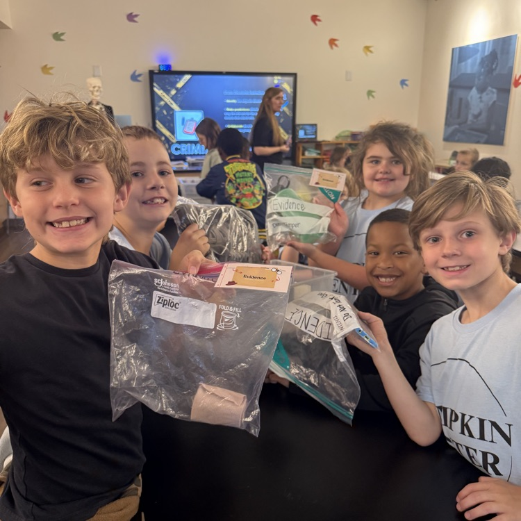 Five kids in a classroom hold clear plastic bags with items. A monitor displays a screen.