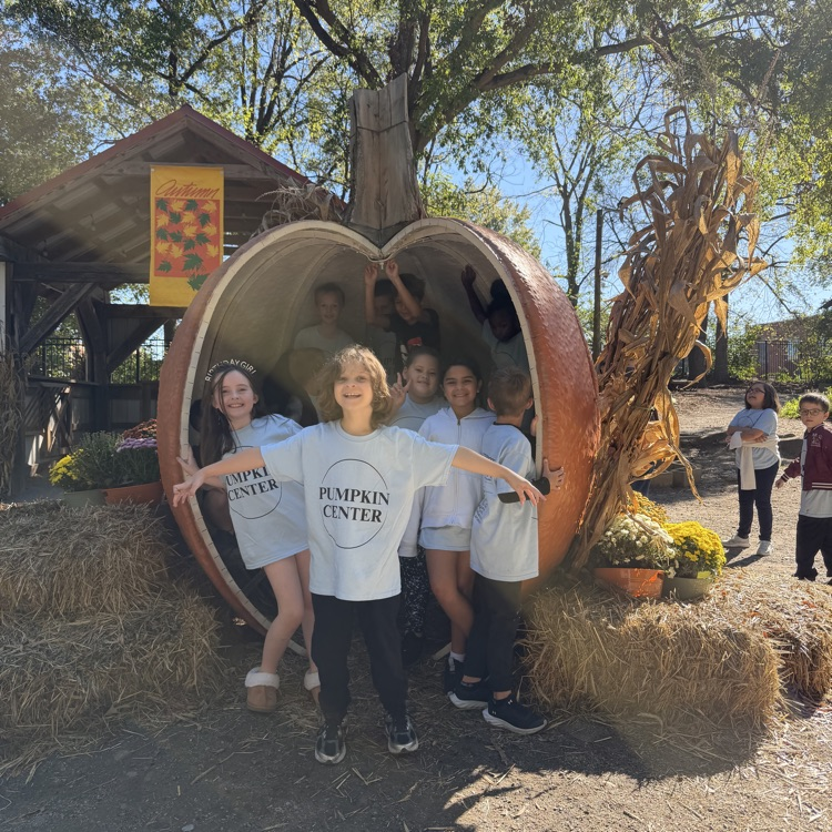 A group of children stand inside a pumpkin and pose for a picture. Hay bales and plants are behind them.