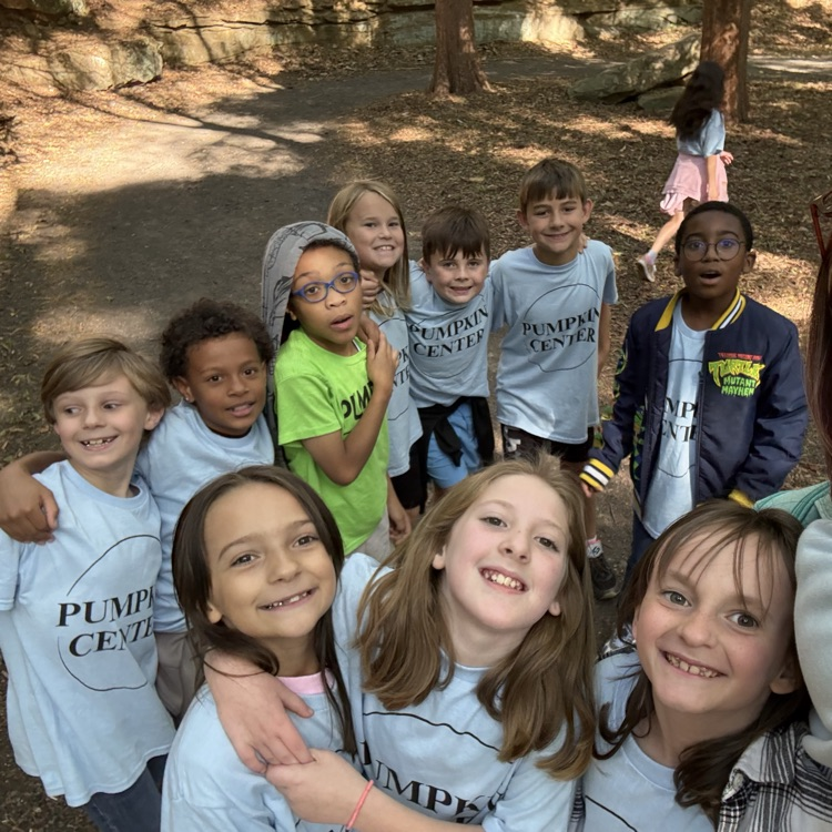 Kids in outdoor area, several wearing white shirts with black letters, one in green, all smiling.