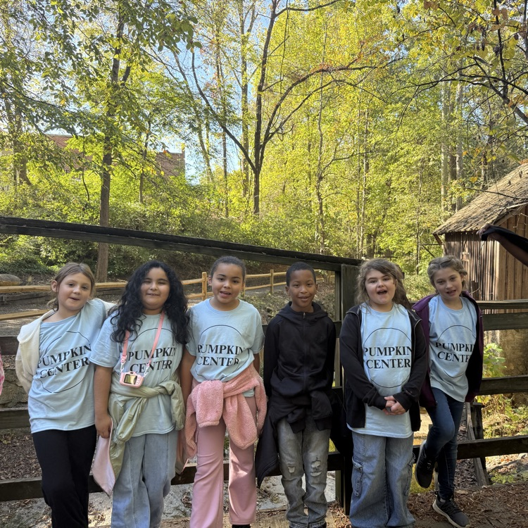 Students in matching shirts stand near a fence, with a wooden building and forest in the background.