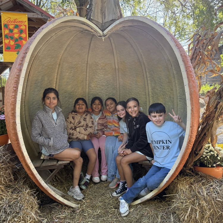 Children pose for photo in large pumpkin. One child holds hand up. Hay in background.
