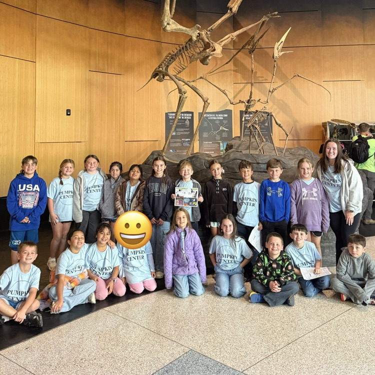 Group of children in various clothes poses in front of a dinosaur skeleton inside a building.