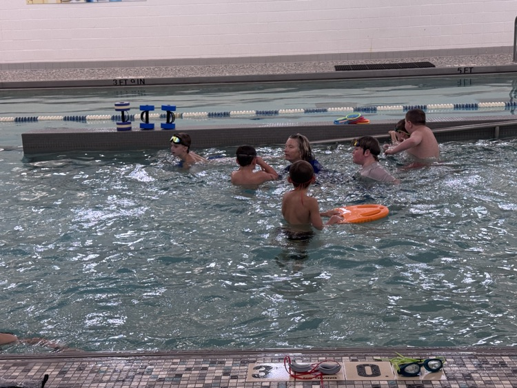 Several young people swimming in a pool with a teacher. A swimmer holds a yellow floatation device.
