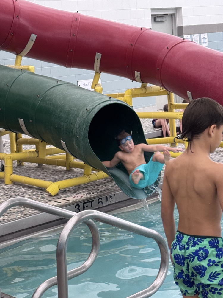 A boy slides down a water slide into a pool, while another boy watches nearby.