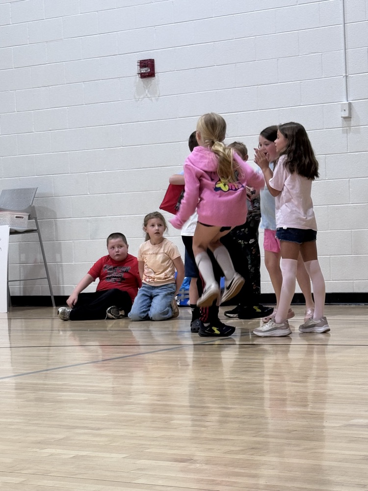 A group of children are gathered in a gym with a wooden floor. Some are sitting while others are standing.