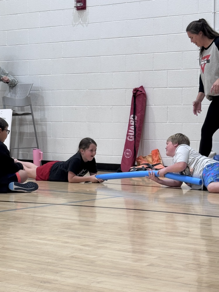 Three children lying on gym floor, each holding a blue tube. One person stands behind them.