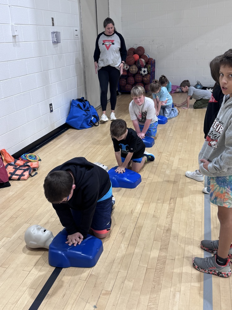 Kids practicing CPR on blue dummies on a gym floor. A teacher stands in the background.