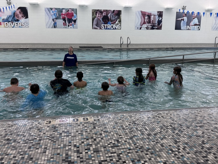 Several children and one adult swimming in a pool while a teacher stands in the water.