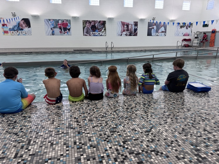 Kids sit on the edge of an indoor pool with tile flooring and pictures on the wall.