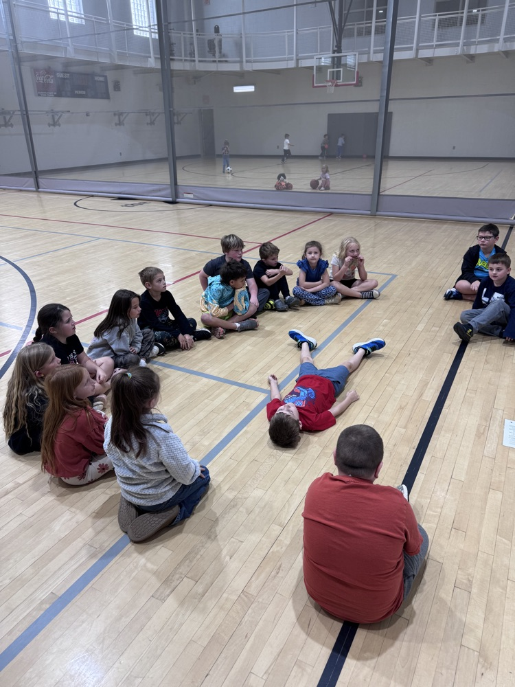 Children sit in a circle on a gym floor; one child lies on the floor while another sits on the edge of the circle.