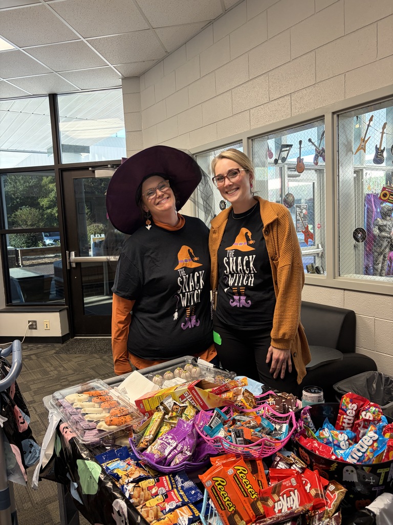 Two women wearing Halloween hats pose behind a table with a large variety of candy.