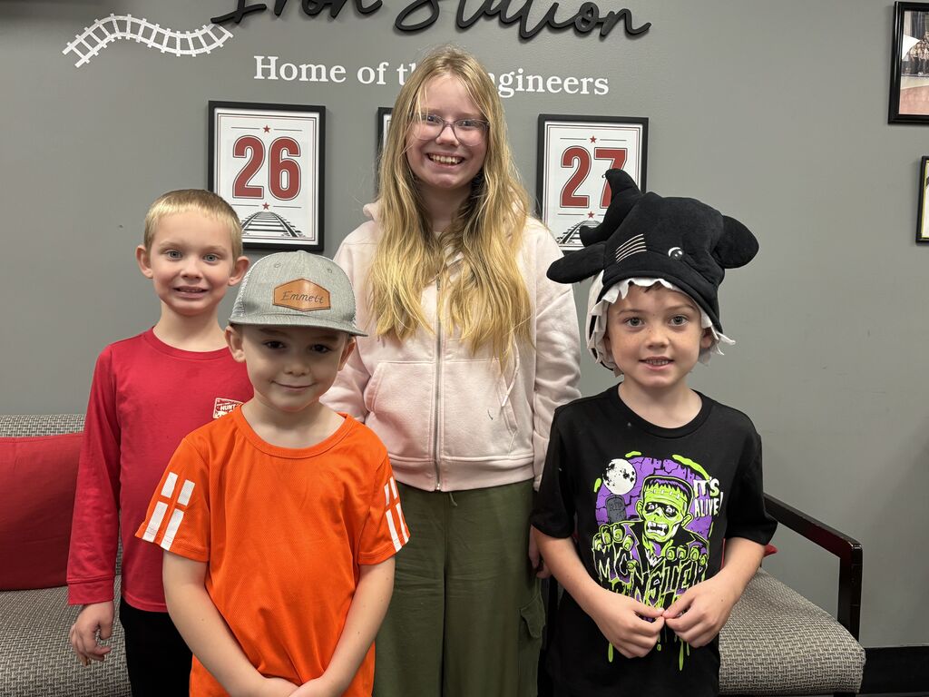 A female student with glasses standing behind three young male students. The boy on the left wears a long-sleeve red shirt. The boy in the center wears a bright orange shirt and a grey baseball hat. The boy on the right wears a black shirt featuring a Frankenstein graphic and a black shark hat. On the wall in the background, a sign reads "Iron Station Home of the Engineers" above framed prints displaying numbers 26 and 27.