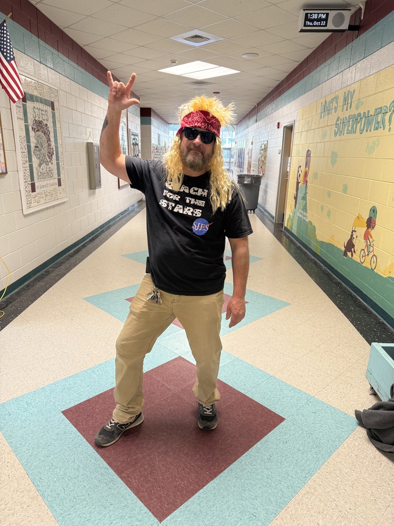 A man in a hallway with long blonde hair, sunglasses, and a bandana poses for a photo.