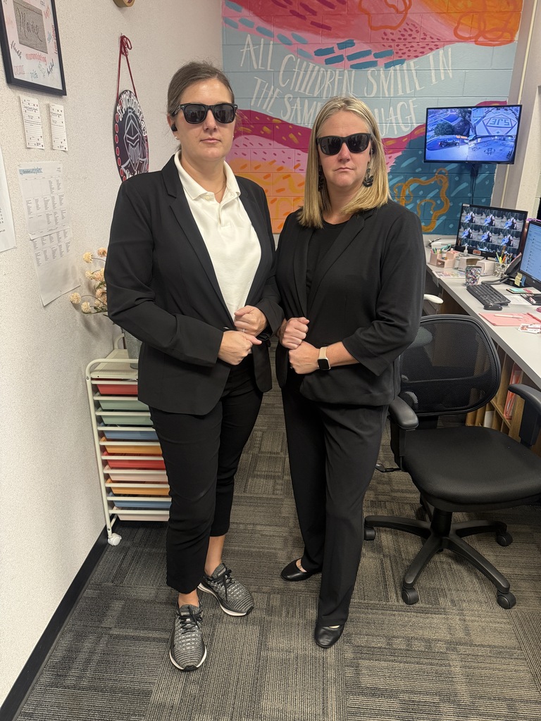 Two women wearing sunglasses and black suits stand in an office with monitors, a desk, and a colorful mural.