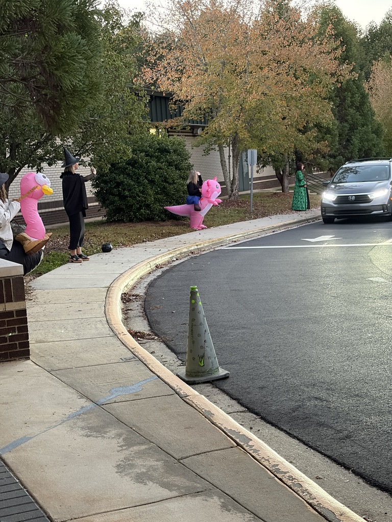 People wearing costumes and sitting on a sidewalk with a traffic cone near a parked car.