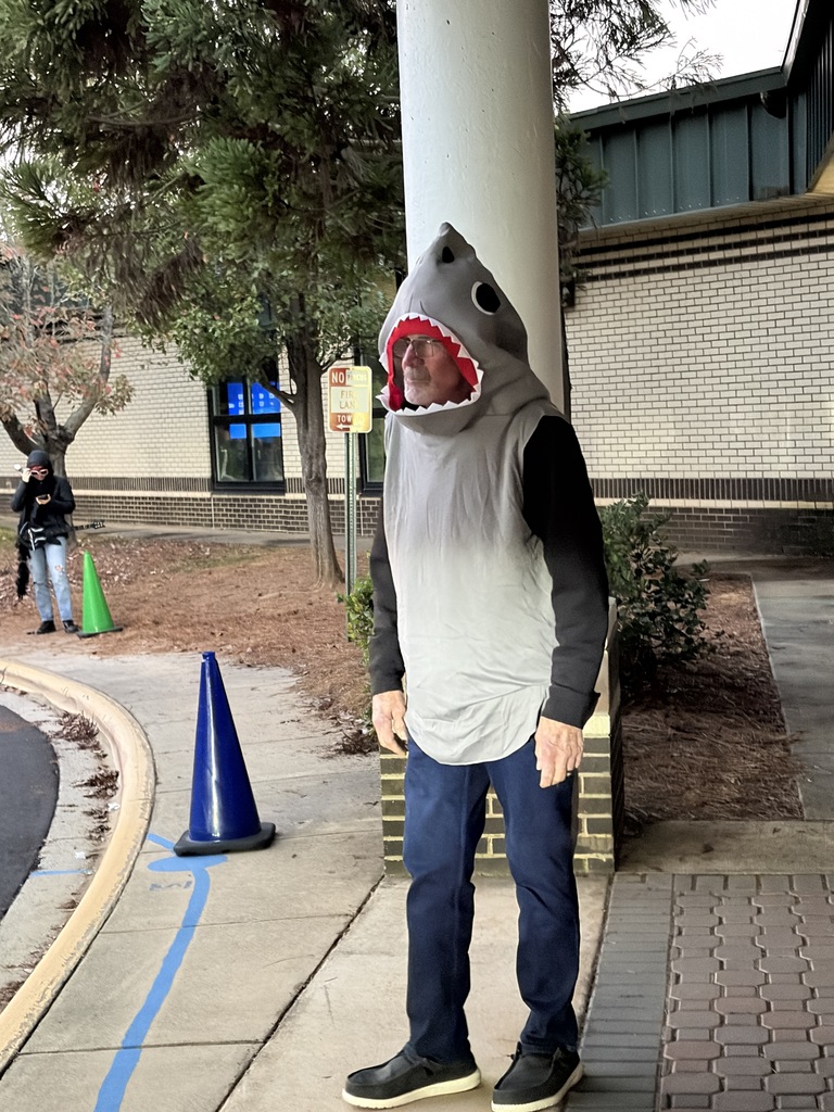 A person in a shark costume stands near a building, wearing a white shirt, blue jeans, and black shoes.