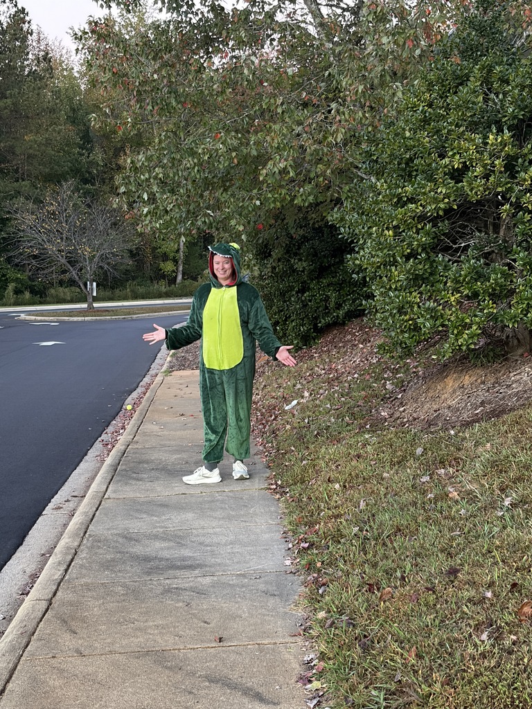 Person in green costume with hood stands on sidewalk with arms outstretched, near a sloped grassy area.