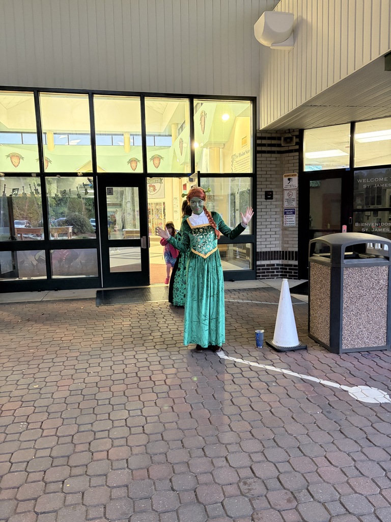 Woman in green costume with red mask stands with arms raised in front of trash can.