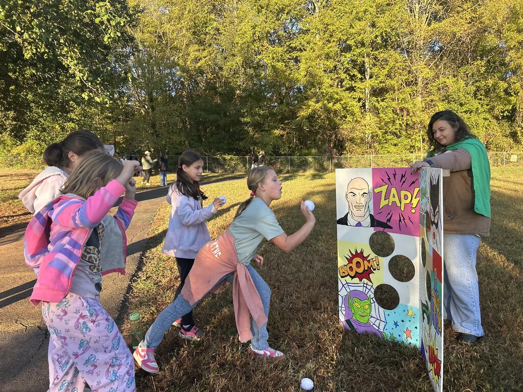 Alt-Text: Four young female students participate in an outdoor carnival game on a sunny field. One student winds up to throw a white ball toward a superhero-themed target board featuring comic art cutouts and target holes. An adult wearing a green cape stands behind the target board helping supervise the activity.