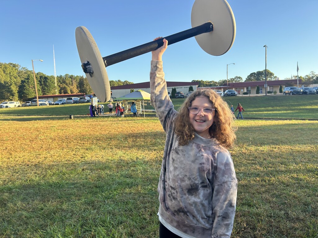A smiling young female student with glasses and curly hair stands on a grassy field outdoors holding a lightweight, oversized prop barbell above her head with one hand. In the background, school buildings, parking lots, and an outdoor canopy tent are visible under a clear, bright sky.
