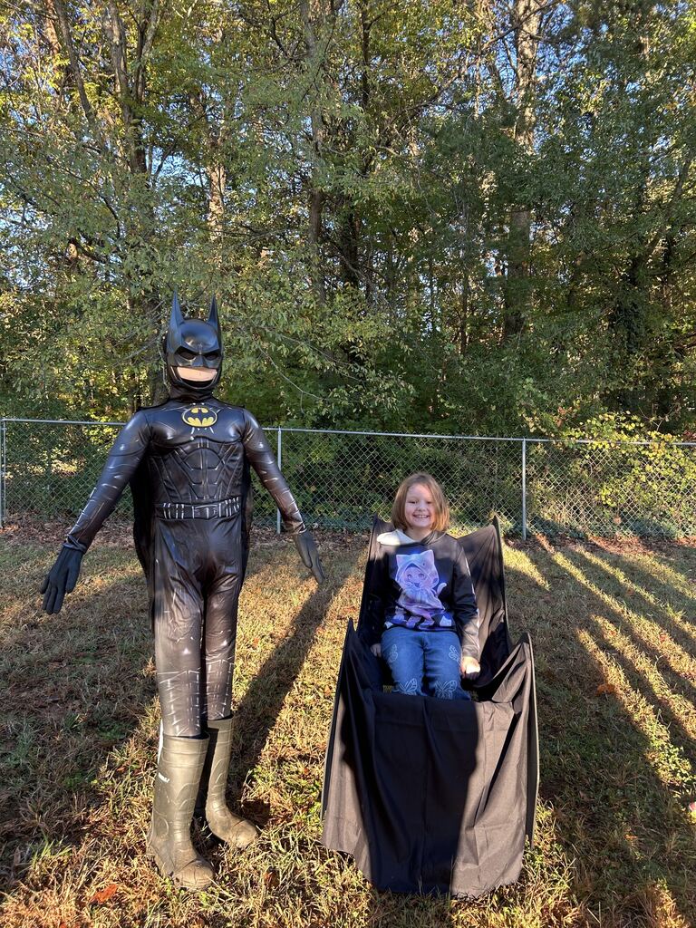 A Batman statue stands outdoors on a grassy lawn next to a young child seated in a chair draped with black Batmobile wings or a black cape. In the background, there is a chain-link fence backed by green and autumn trees illuminated by sunlight.
