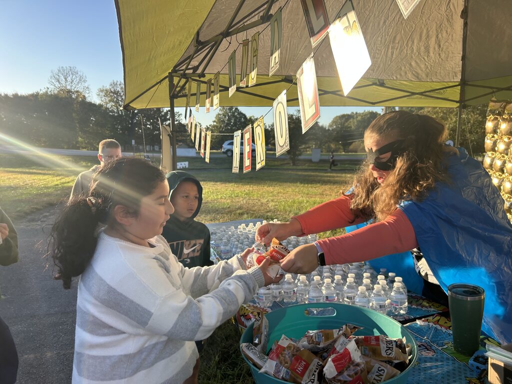 An adult wearing a black superhero mask and a blue cape hands a bag of snacks to a young female student at an outdoor tent station. Table displays show bottled water, snack bags in a tub, and a banner hanging above reading "DIG IN SUPERHEROES." Other young students stand nearby waiting in line outdoors under bright sunlight.