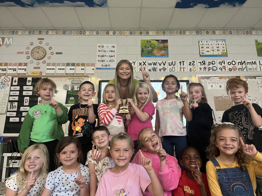 Students standing in front of a teacher. One student poses with a trophy. One student gives a peace sign.