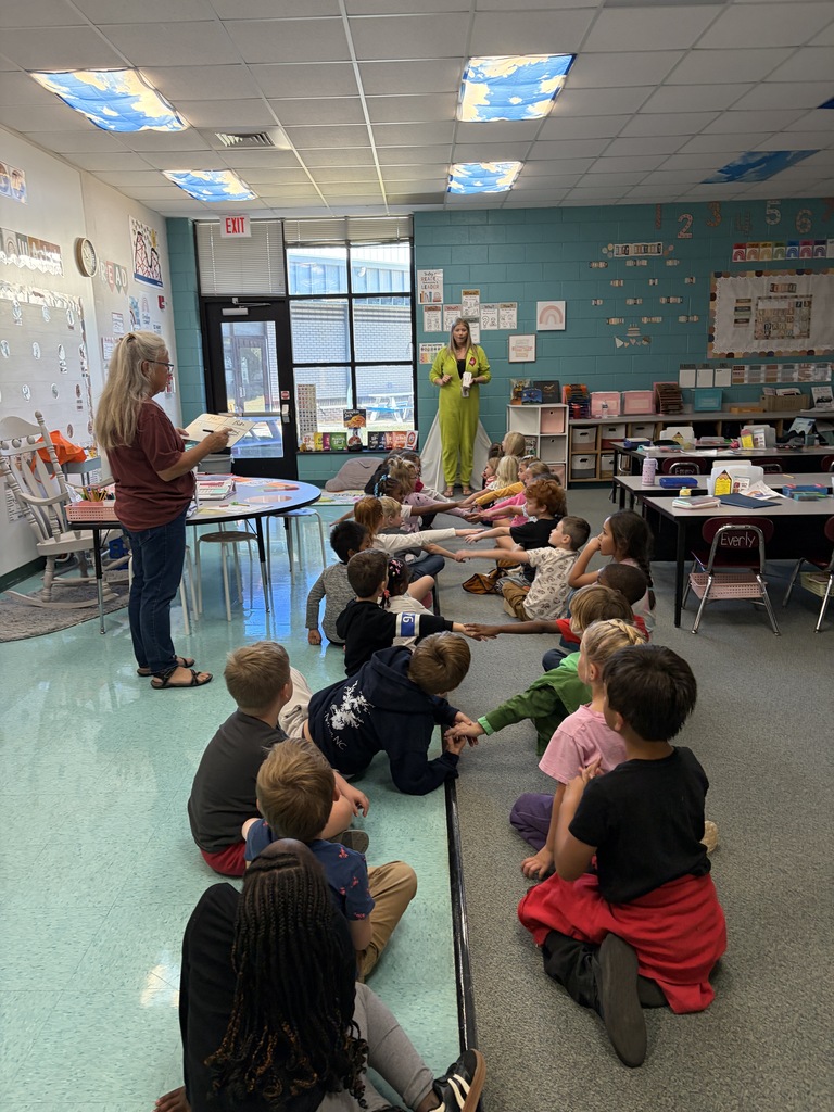 A classroom with children seated on the floor. A woman stands before them reading from a book.