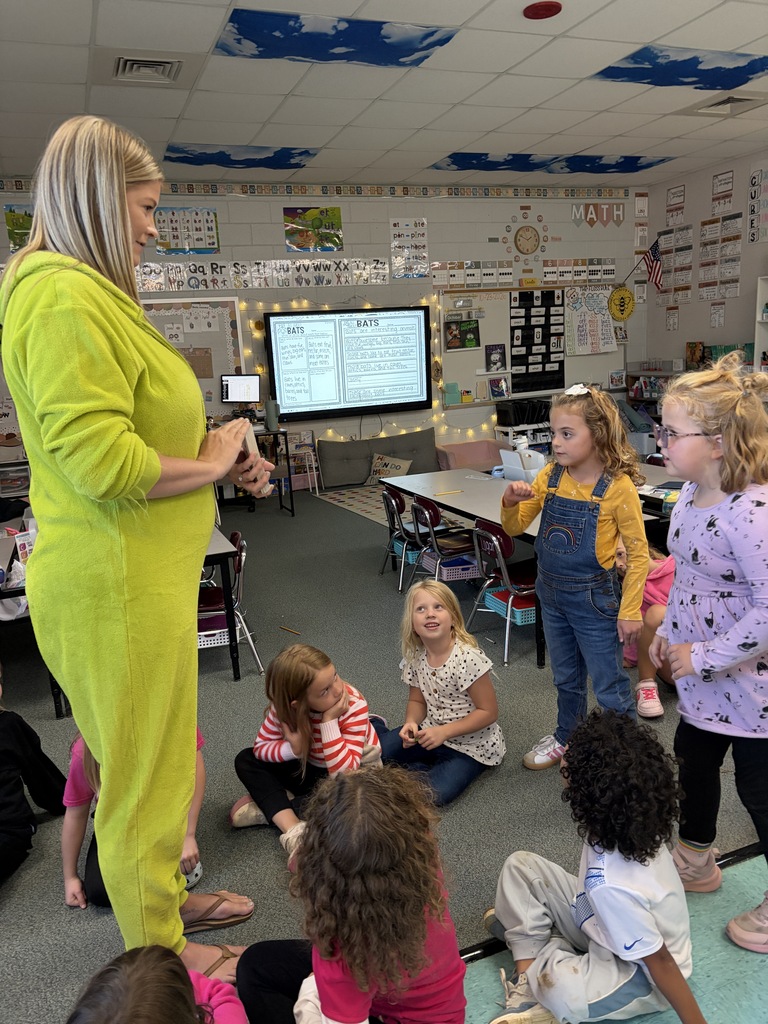 Woman in yellow jumpsuit speaks to young children seated in circle, two hold books; room has tables, chairs, screen.