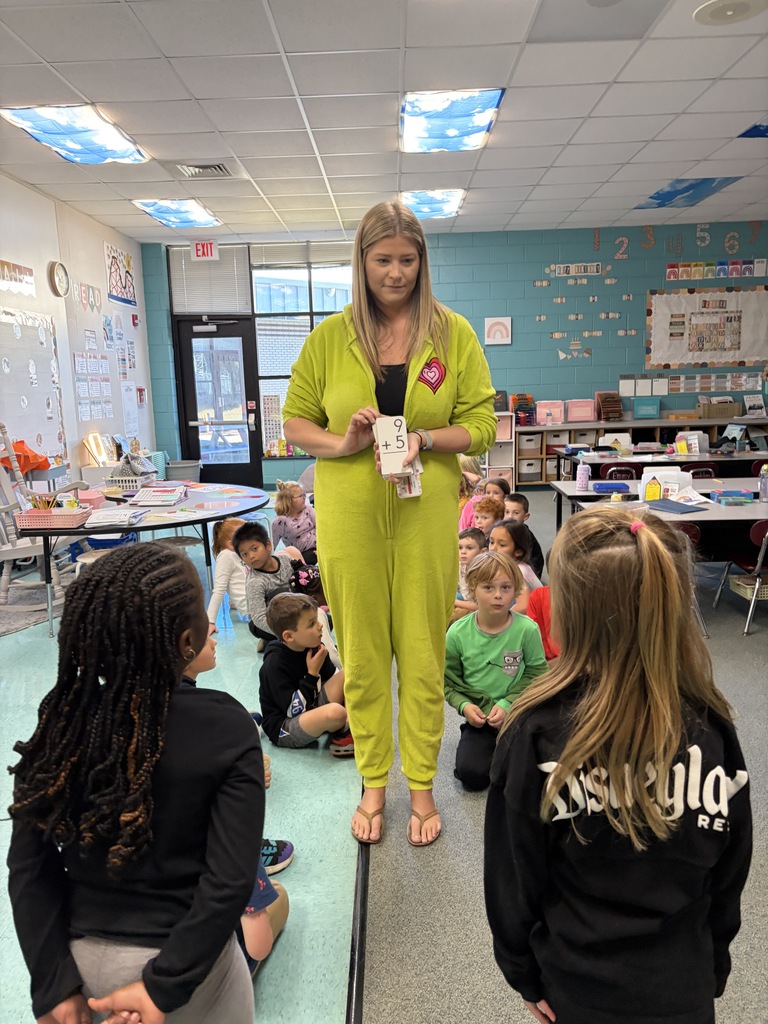 A teacher in green jumpsuit stands in front of children seated in a circle, holding a book.