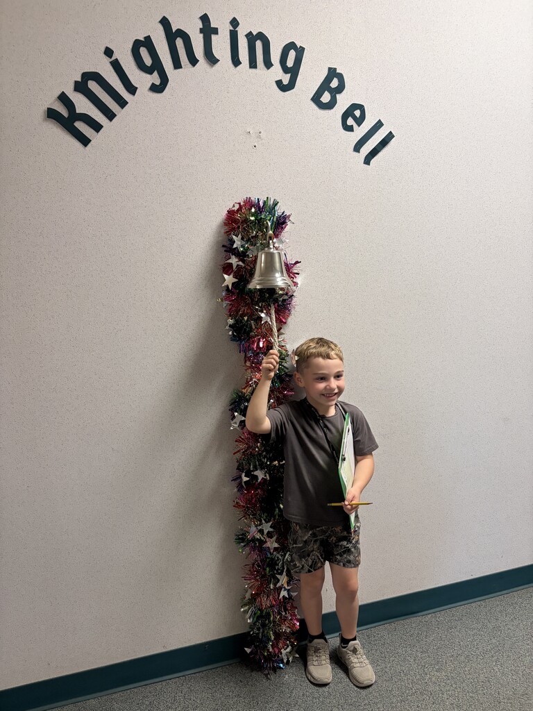 Boy holding bell and ribbon next to a decorated wall with the words "Knighting Bell".