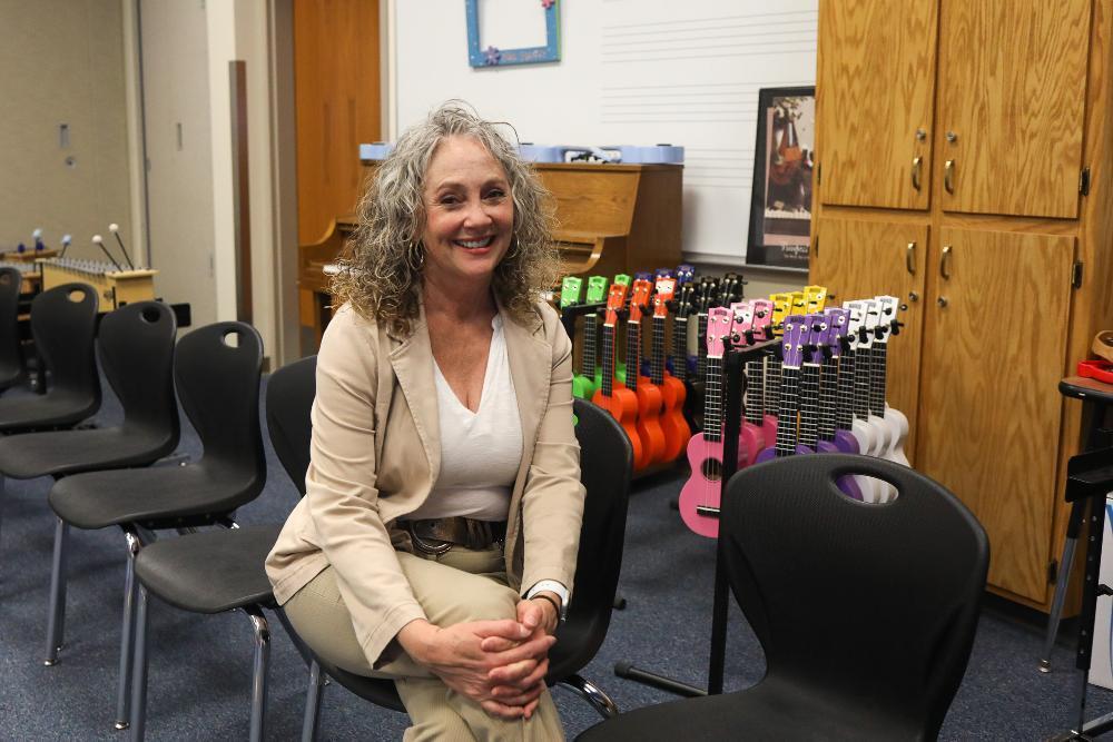 A smiling woman with curly gray hair and a beige jacket sits in a music classroom, looking at the camera. Behind her are rows of colorful ukuleles, a piano, and more black chairs.