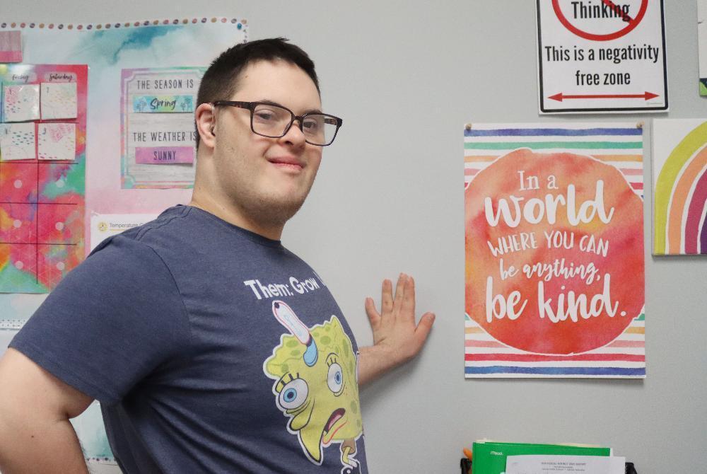 A smiling young man with glasses wears a blue Spongebob shirt. He stands in a classroom, his right hand touching a wall next to a colorful "Be Kind" poster. Educational charts are visible behind him.