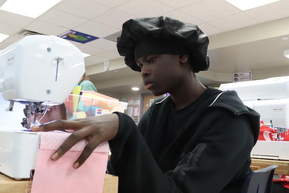A young man, wearing a black chef's hat and hoodie, intently sews pink fabric on a white sewing machine. His hands carefully guide the material, focused on the task in a bright classroom setting.