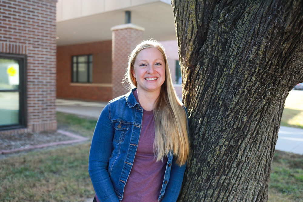 A smiling woman with long blonde hair wearing a denim jacket and purple shirt stands beside a large tree outside a brick school building.
