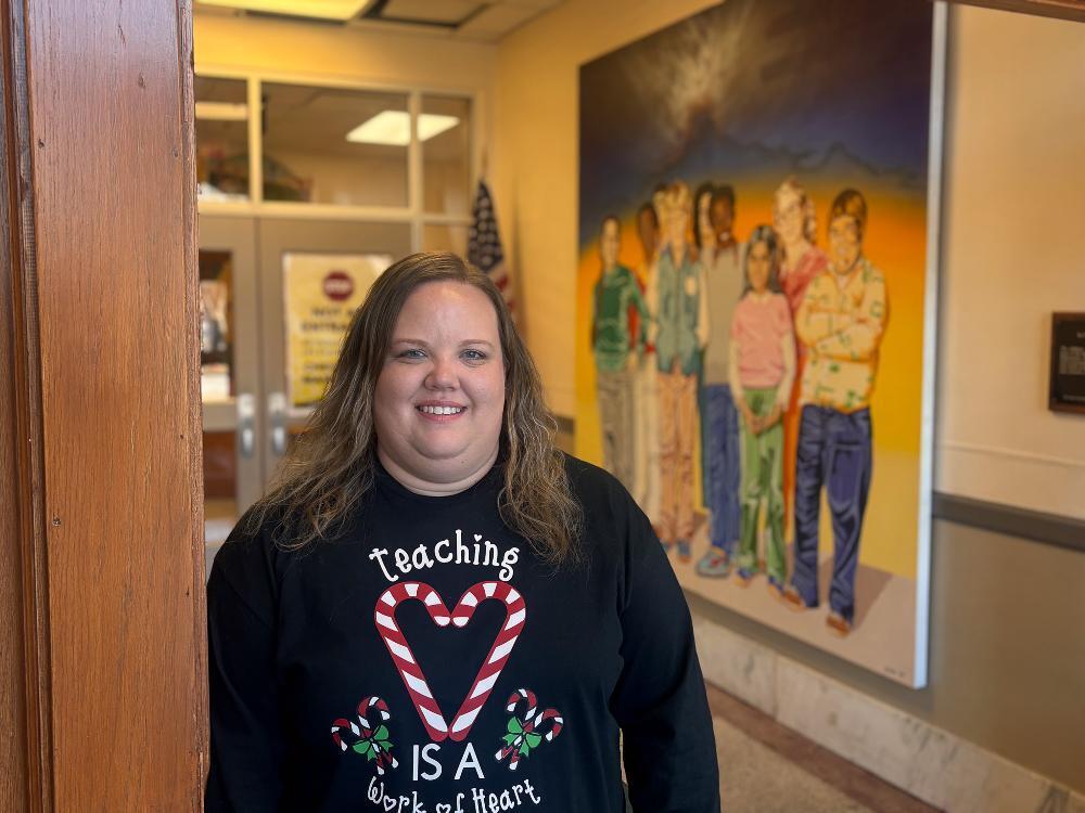 A smiling woman in a holiday "Teaching is a work of heart" shirt stands in a school hallway with a student mural behind her.