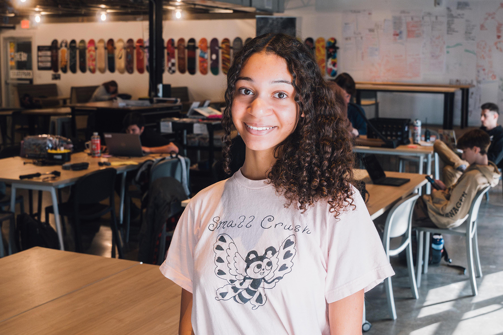 Bay High Focus Program senior Avery smiles while standing in the main Bay High classroom area. Several students are sitting at desks behind her.