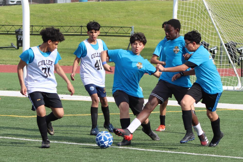 Soccer players, in blue and white uniforms, compete during a match on a green field. "CULLER" and numbers are visible on their jerseys. One player is actively dribbling the ball.