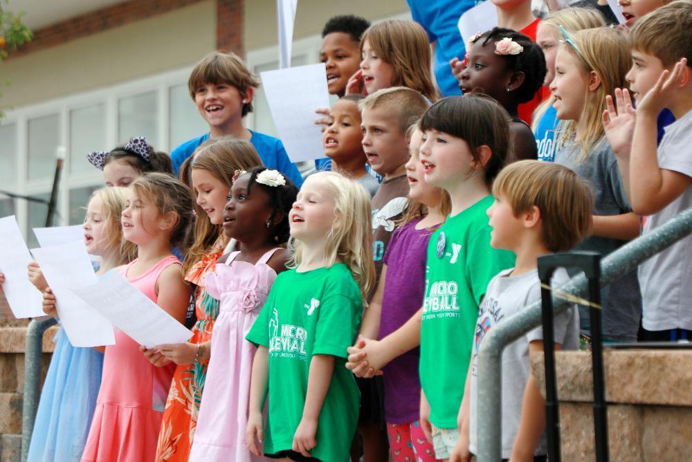 Three rows of Randolph Elementary School students sing on steps outside the building during a festive event this fall. The building will turn 100 years old on Jan. 25, 1926. Several students in the front row are holding sheets of music, and other children are singing the words from memory. They are wearing dresses and t-shirts and are singing loudly toward an audience.