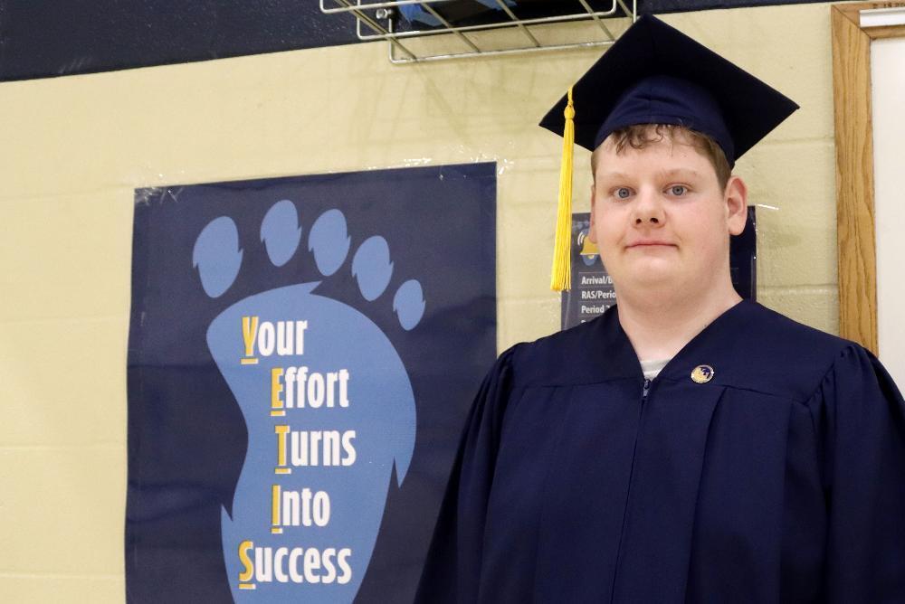 A young graduate in a dark blue cap and gown with a yellow tassel looks forward. Behind him is a blue poster featuring a paw print and the text "Your Effort Turns Into Success."