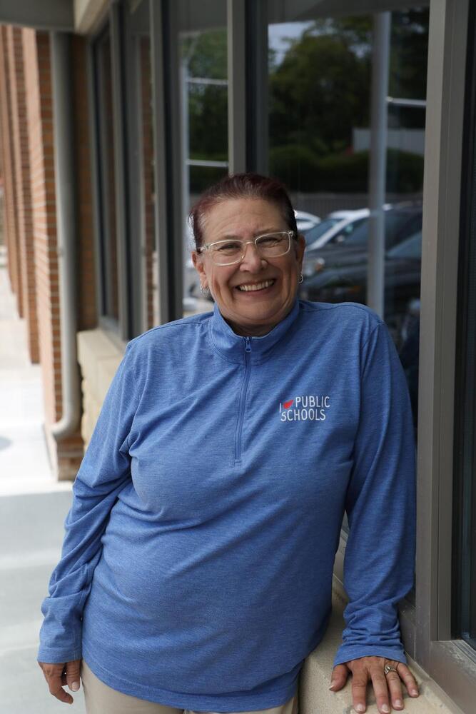 A woman, wearing a blue "I ? PUBLIC SCHOOLS" shirt, smiles while leaning against a brick building's exterior.