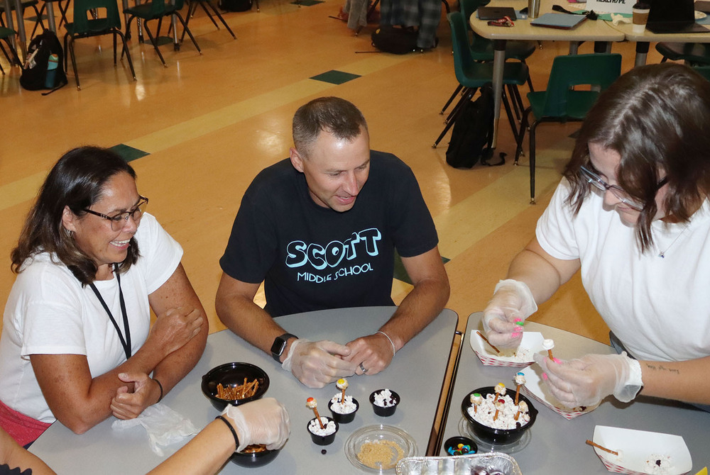 Two Scott Middle School staff members smile as they watch a teammate work on a Scott Chopped activity in the multipurpose room. They are sitting at a table. The person who is working on the activity is standing on the righthand side and is placing a piece of candy on a pretzel stick.